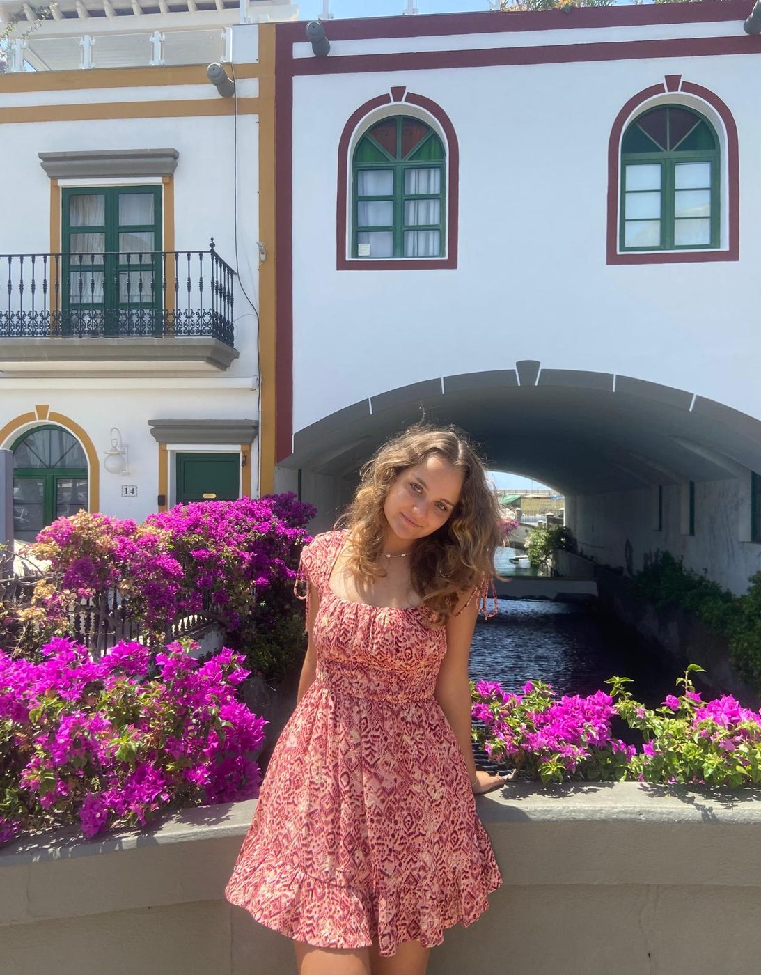 Bianka Kormos in a summer dress, standing in front of a white house with arched green windows and purple bougainvillea