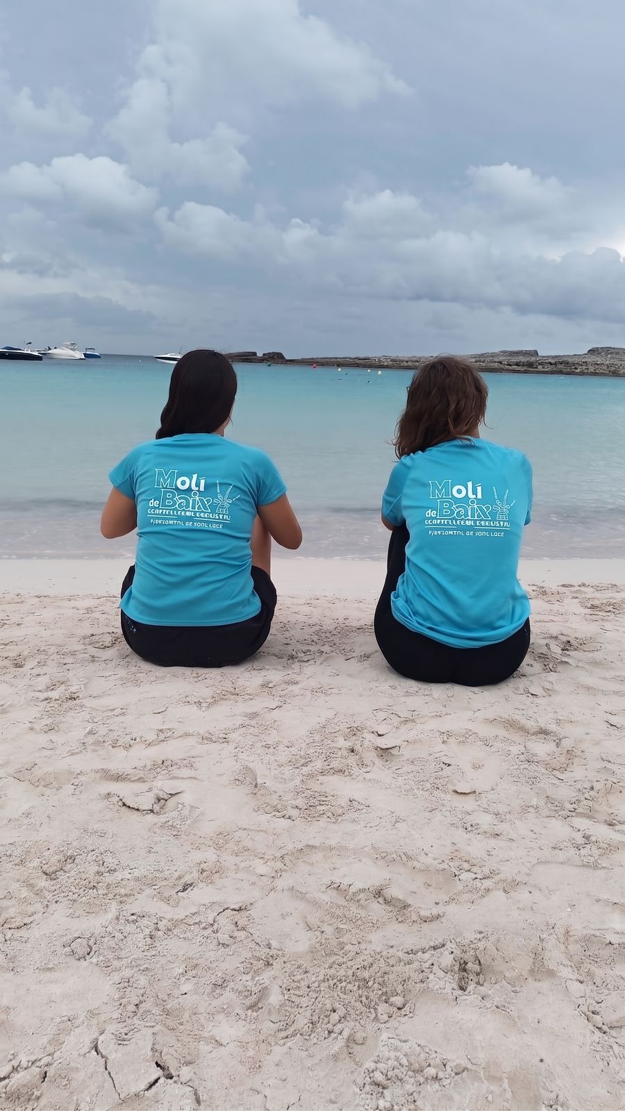 Two volunteers in turquoise Molí de Baix T-shirts sitting on a white sand beach, looking out to sea