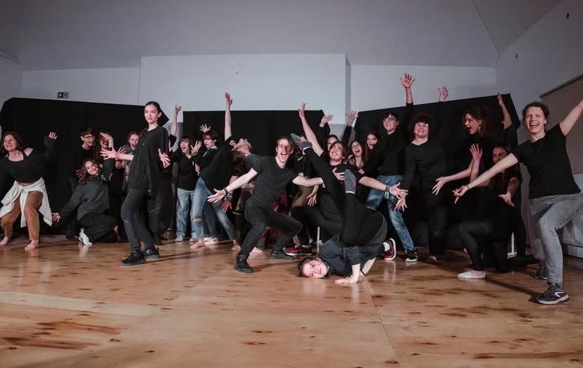 The JESZ student theatre group in black, striking a joyful pose together on a wooden stage floor